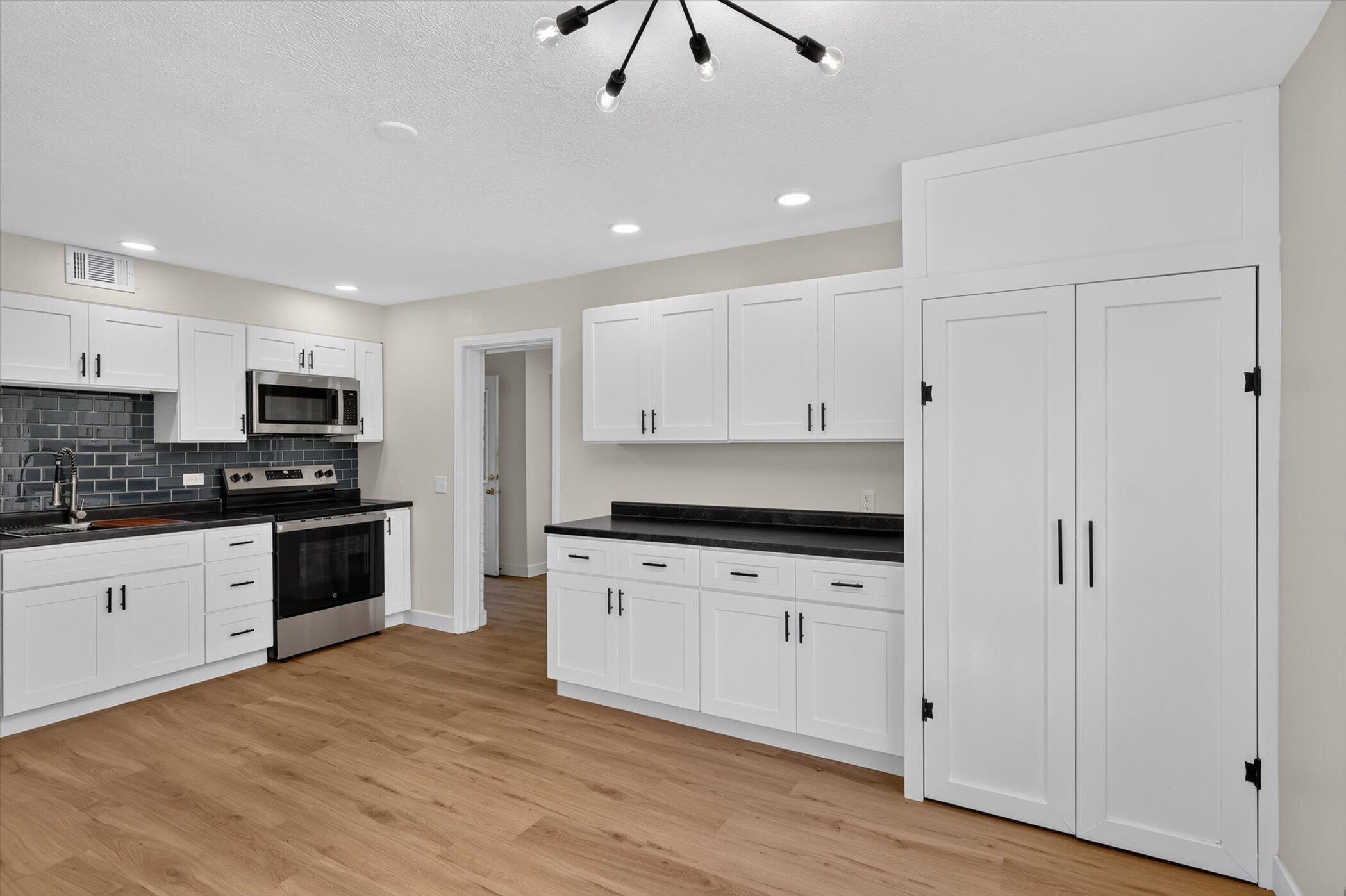 Kitchen with white cabinets, black countertops, dark tile backsplash, stainless steel appliances, and light wood flooring.