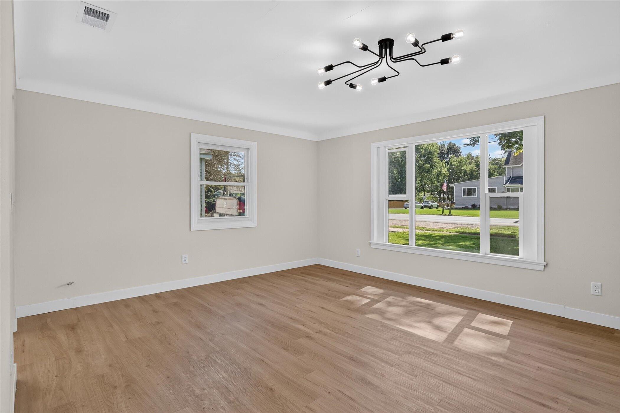 Empty room with light wood floors, a black branching ceiling light, and large windows overlooking a residential street.