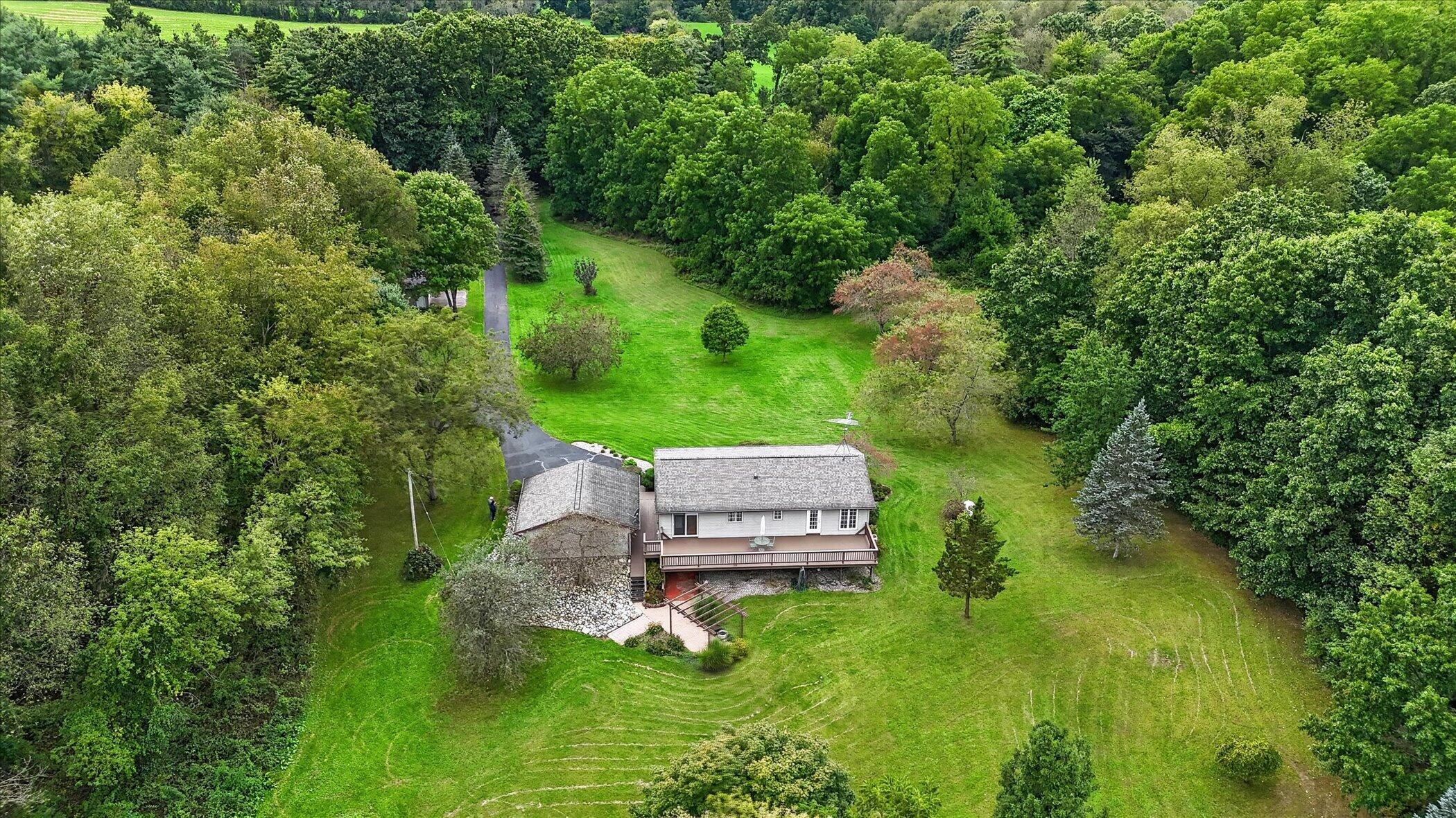 Aerial view of a home with a long deck, paved driveway, expansive lawn, and surrounding mature trees.