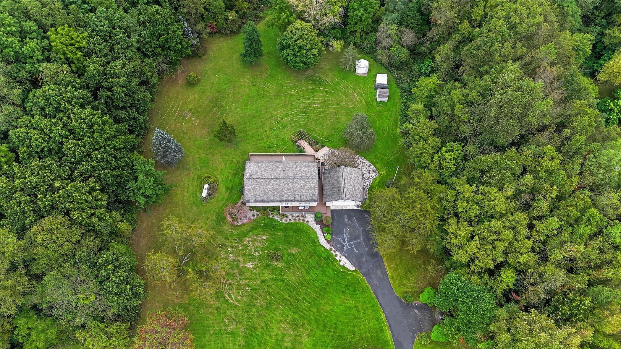 Aerial view of a house surrounded by trees, with a large lawn, driveway, and small outbuildings.