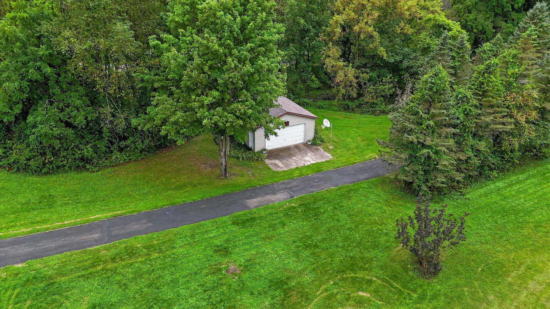 Aerial view of a detached garage beside a paved drive, green lawn, basketball hoop, and surrounding trees.