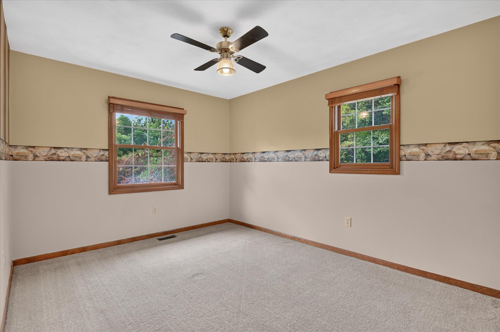 Empty carpeted room with two wood-trimmed windows and a ceiling fan.