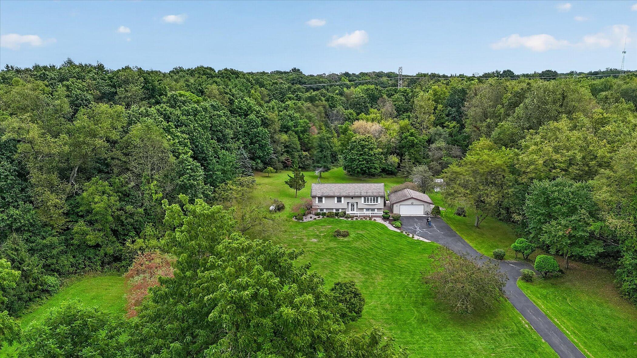 Aerial view of a house and detached garage amid a broad lawn and dense woodland.