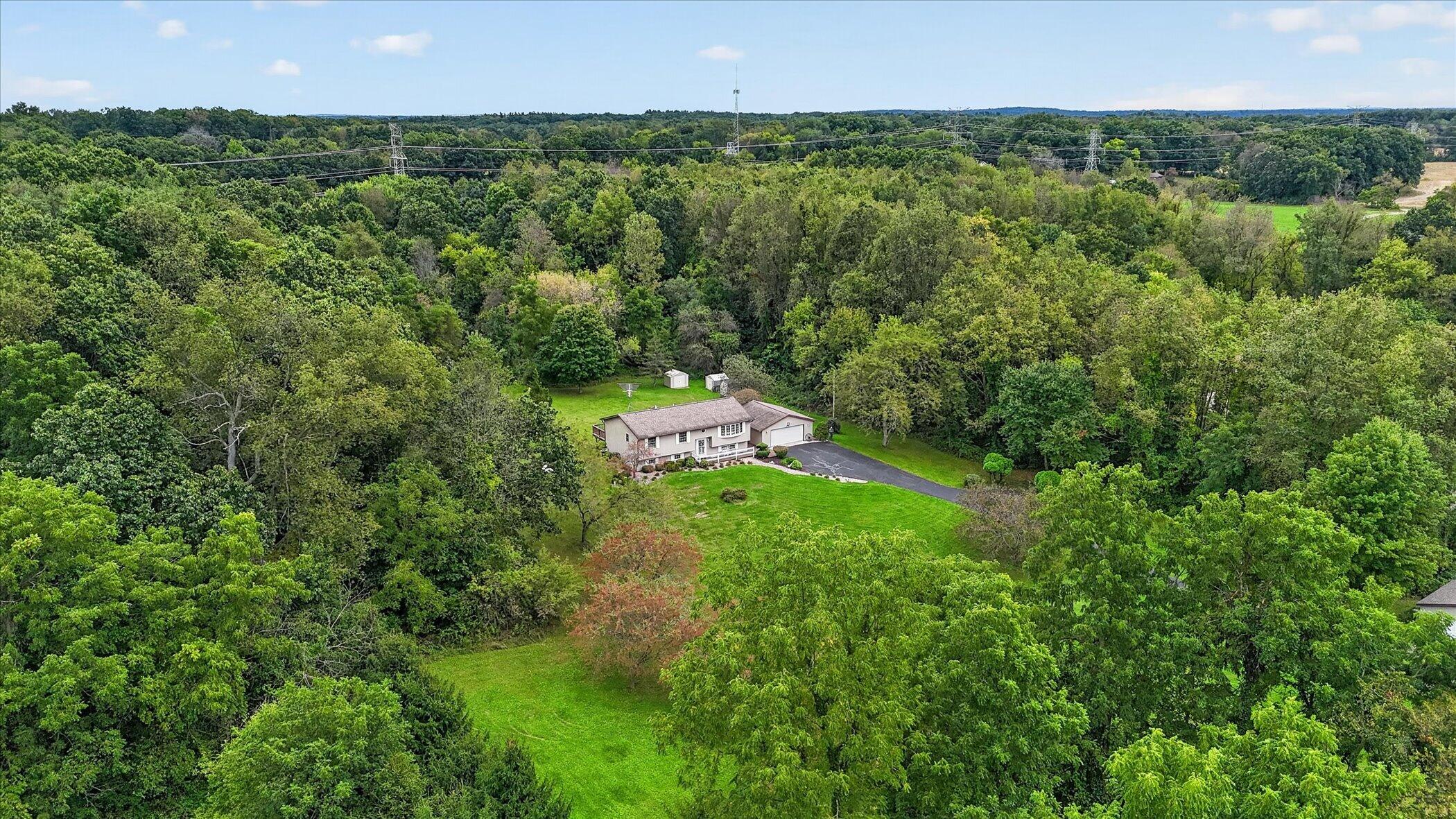 Aerial view of a house in a wooded clearing, with a broad lawn, driveway, and two outbuildings.
