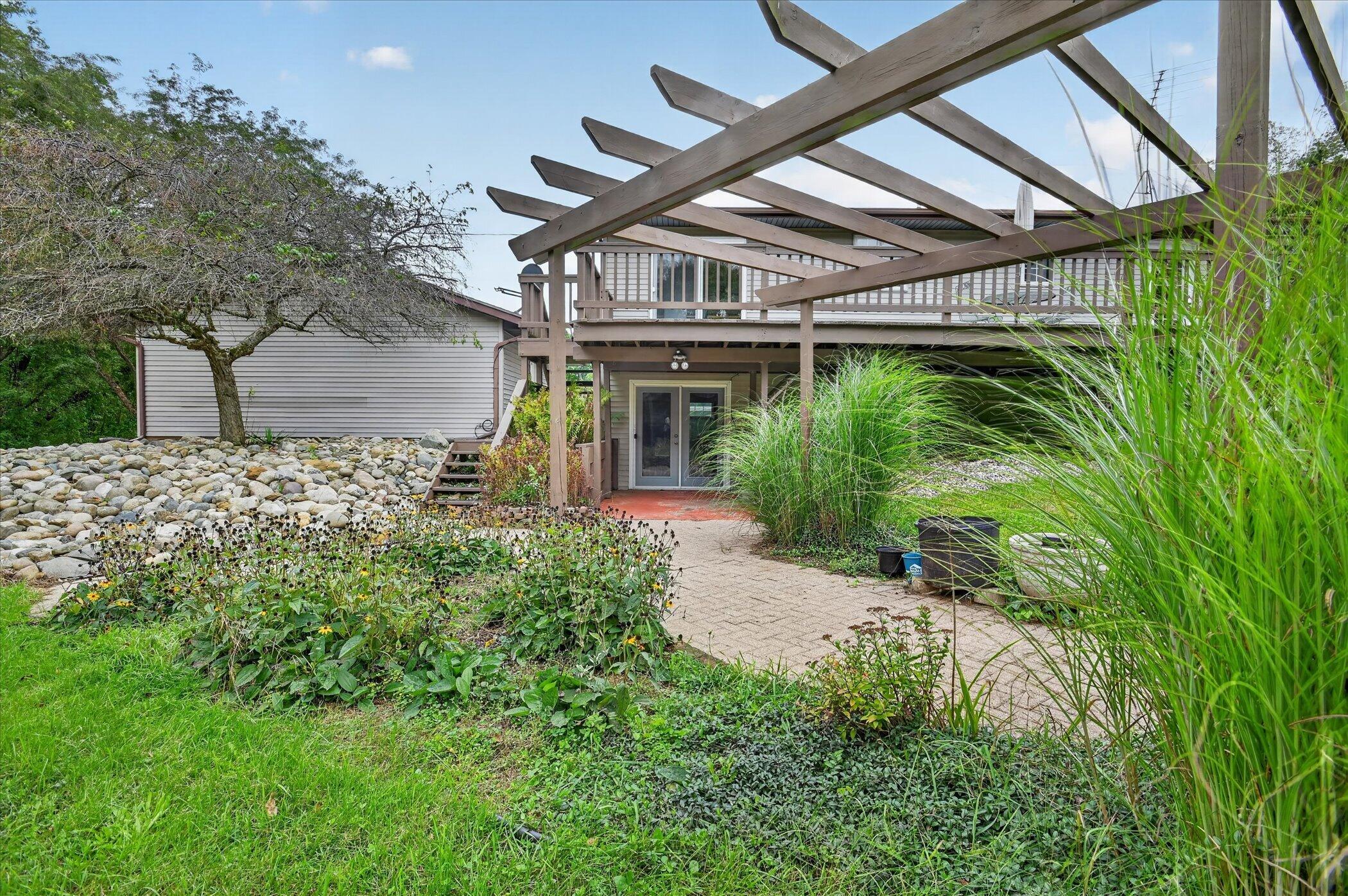 Rear patio with a wooden pergola, raised deck, paved walkway, and ornamental grasses.