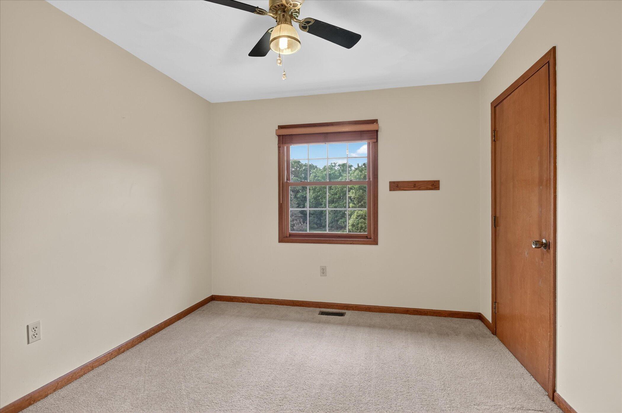 Empty carpeted room with a wood-framed window, ceiling fan, and wooden door.