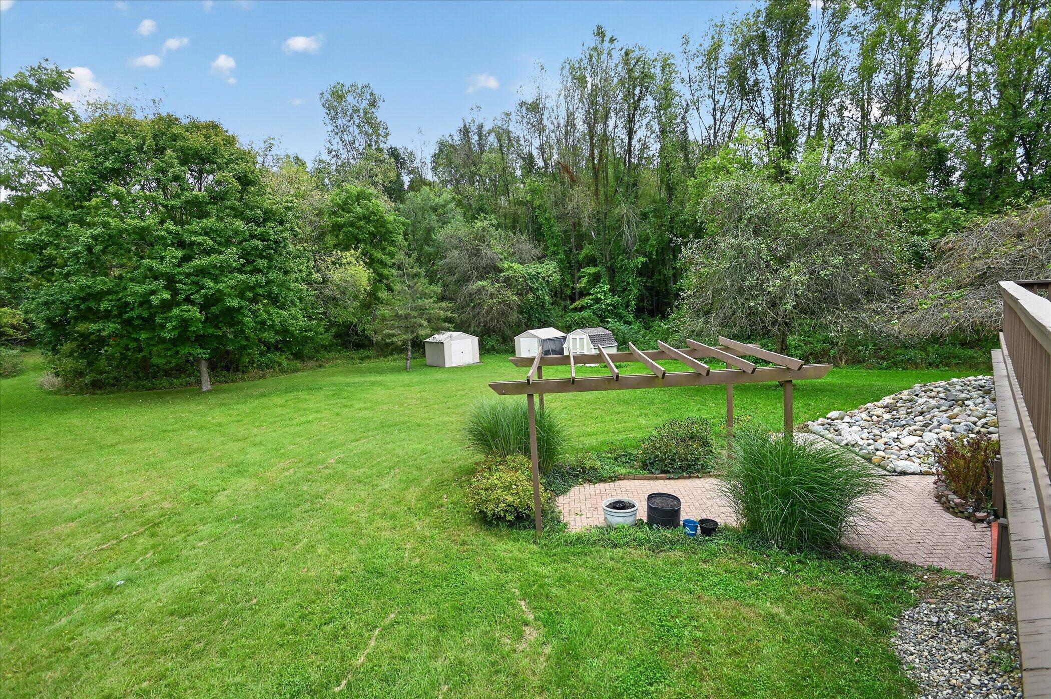 Backyard with a wooden pergola over a brick patio, broad lawn, storage sheds, and wooded backdrop.
