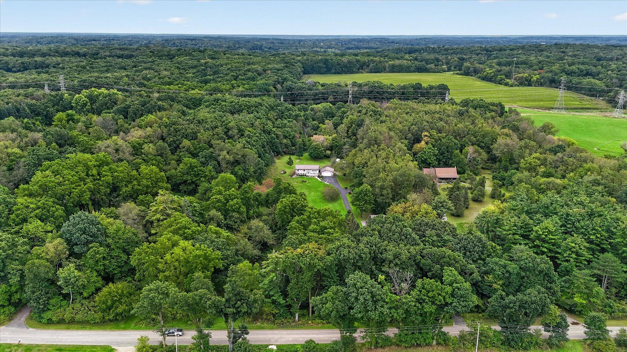 Aerial view of a home and outbuilding amid wooded acreage, open fields, and power lines.