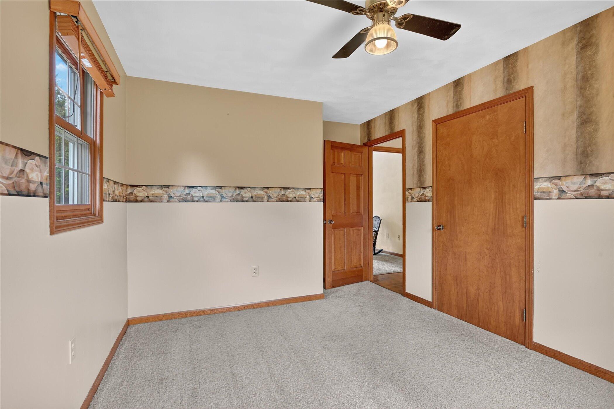 Empty carpeted bedroom with a ceiling fan, wood-trimmed window, and two wooden doors.