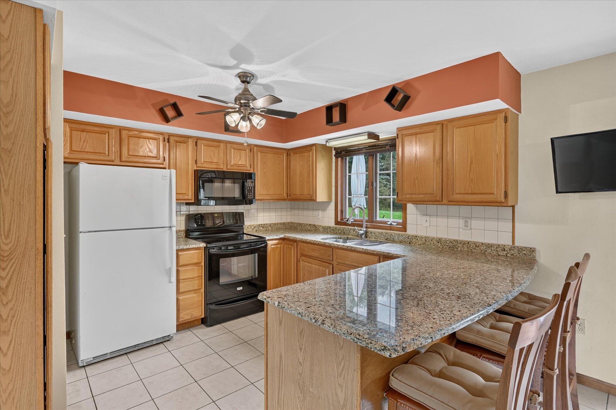 Kitchen with honey-colored cabinets, granite breakfast bar, and stools beside a window over the sink.