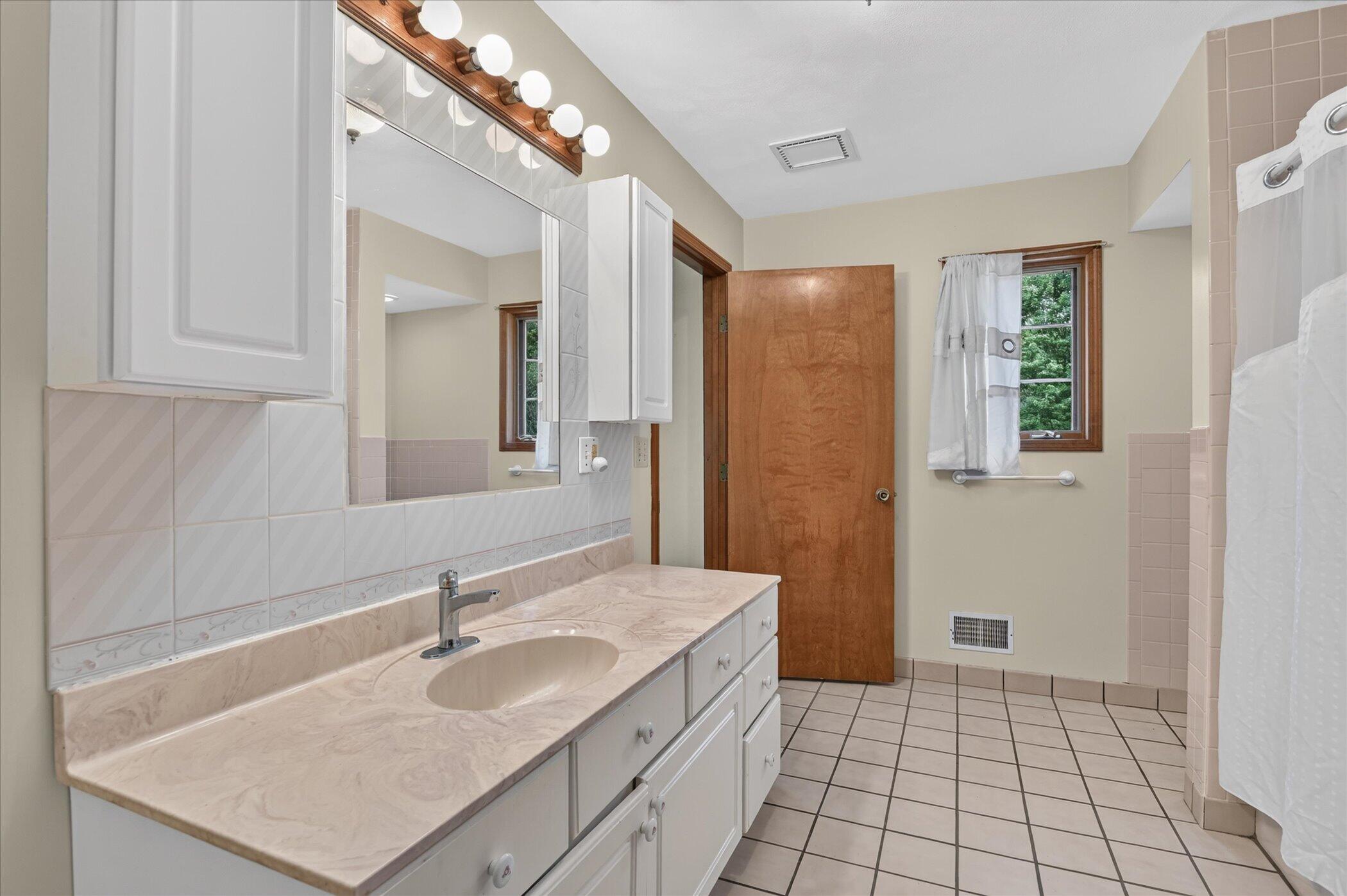 Bathroom with a long white vanity, globe-lit mirror, tiled floor and curtained shower.