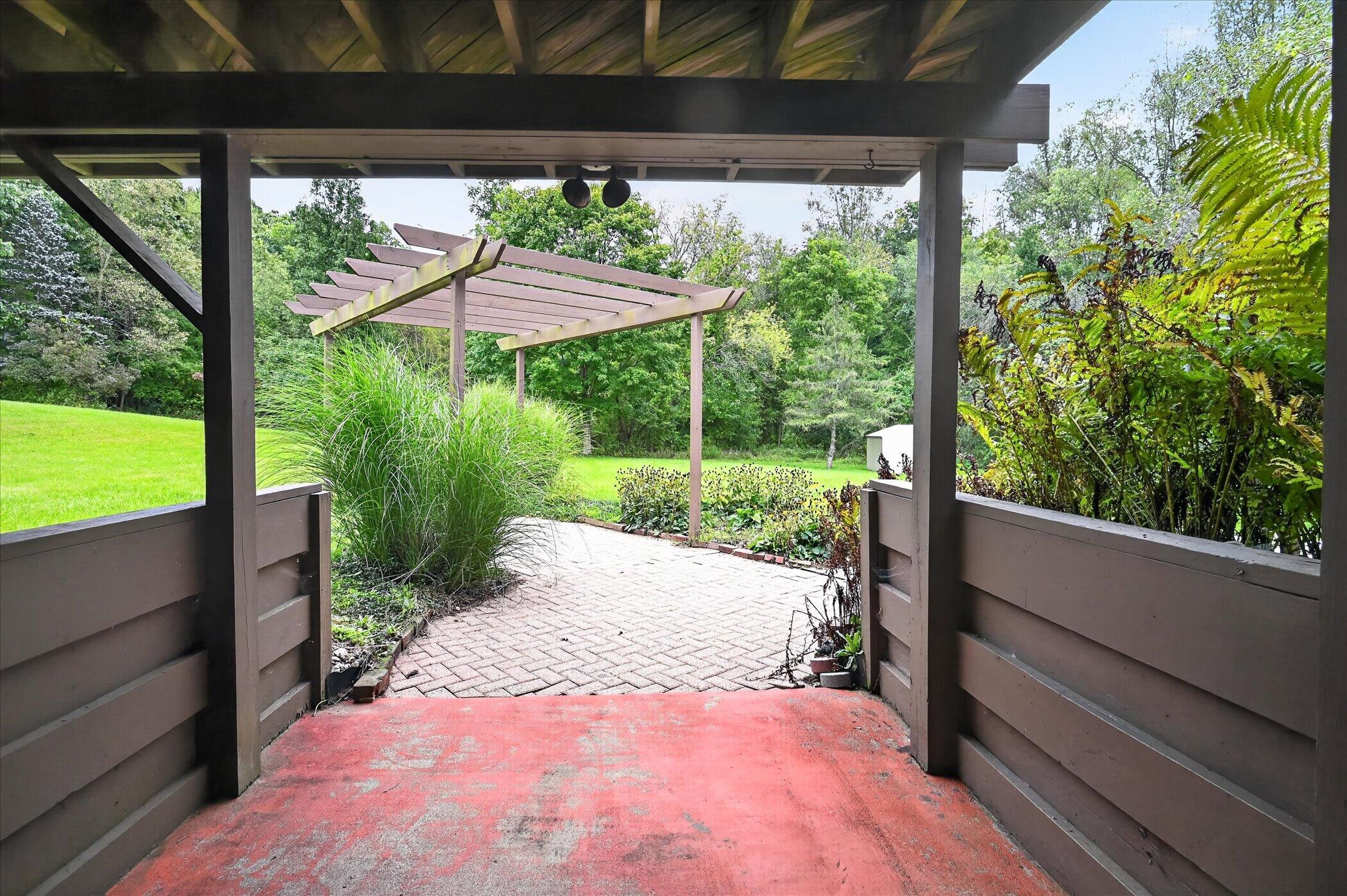 Covered patio overlooking a brick walkway, wooden pergola, landscaped garden, and lawn.