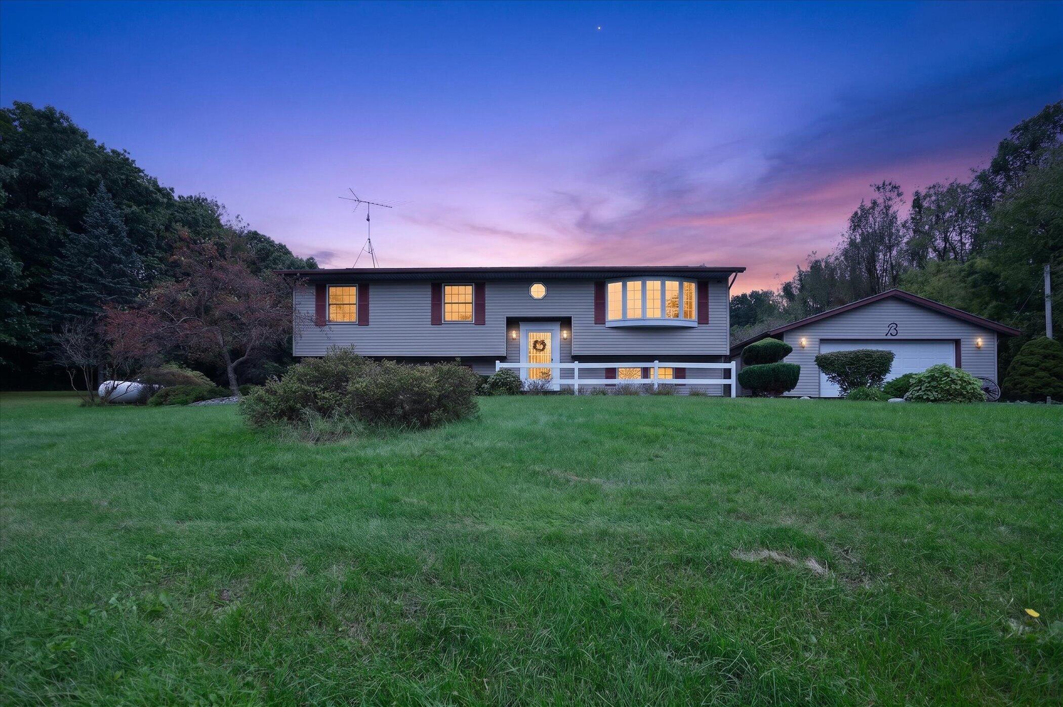 Raised ranch home with illuminated windows, detached garage, and broad green lawn at dusk.
