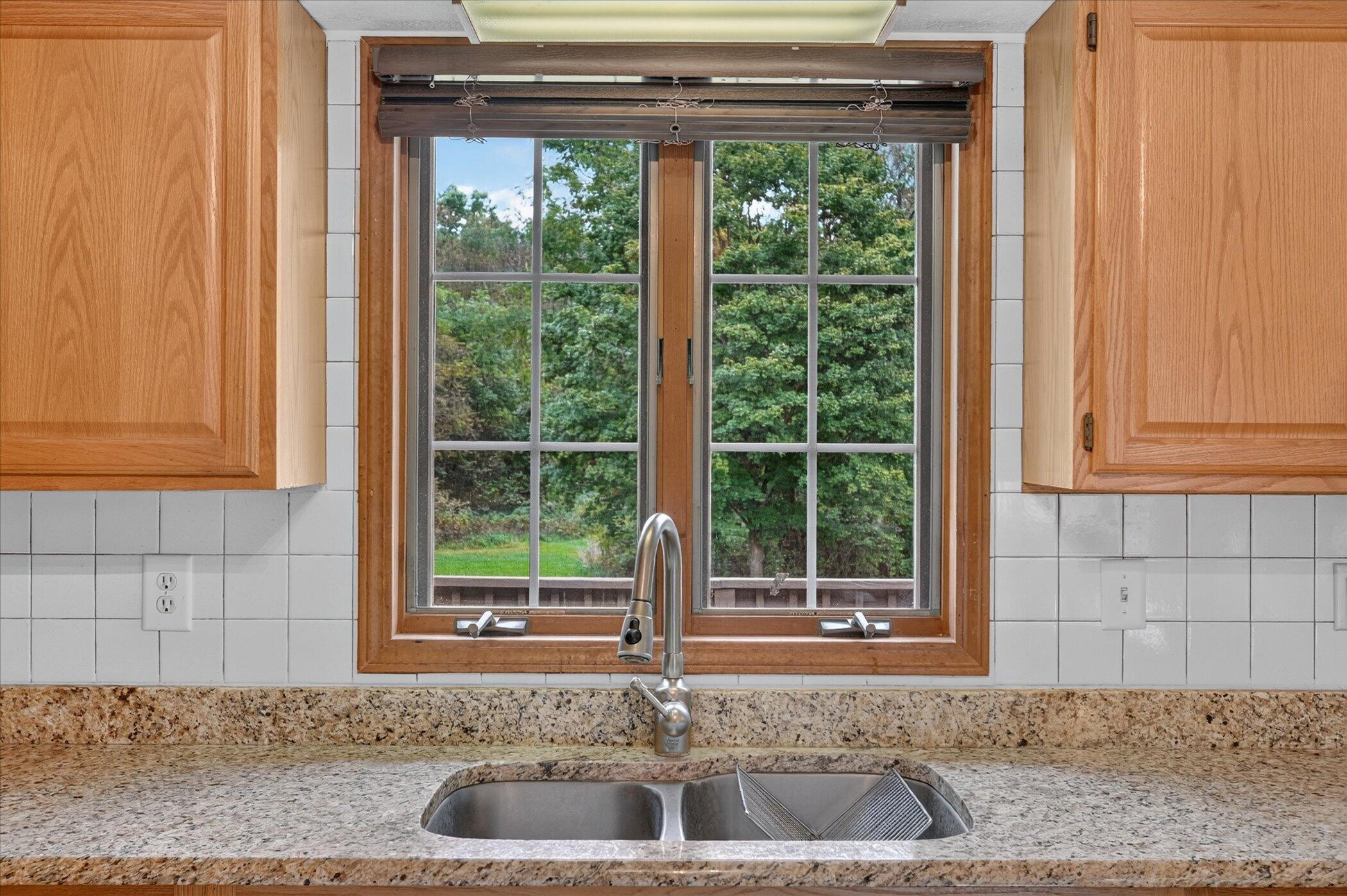 Kitchen sink beneath a wood-trimmed window overlooking trees, set in granite counters and white tile.