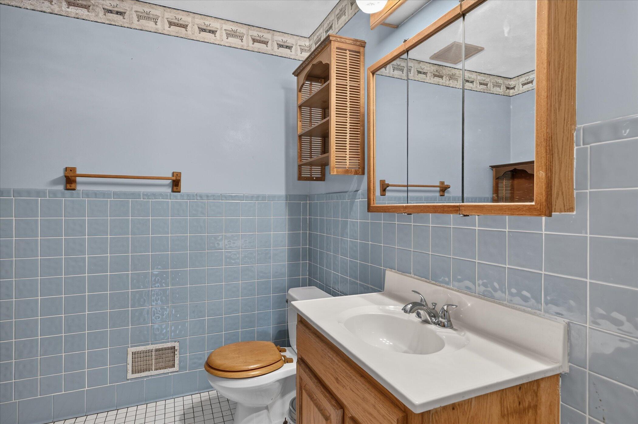 Bathroom with blue tiled walls, a wood vanity and mirrored cabinet, and a toilet with a wooden seat.