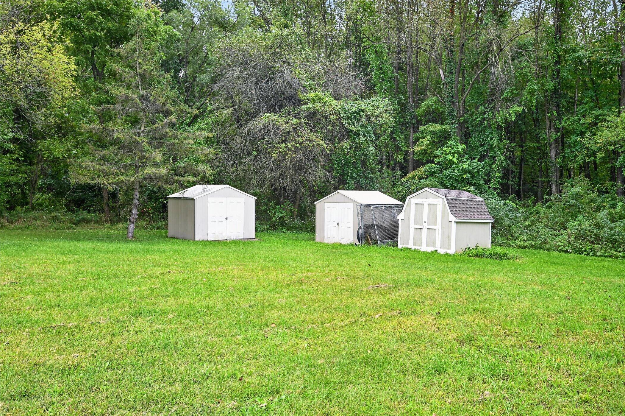 Grassy yard with three white storage sheds backed by dense woods.