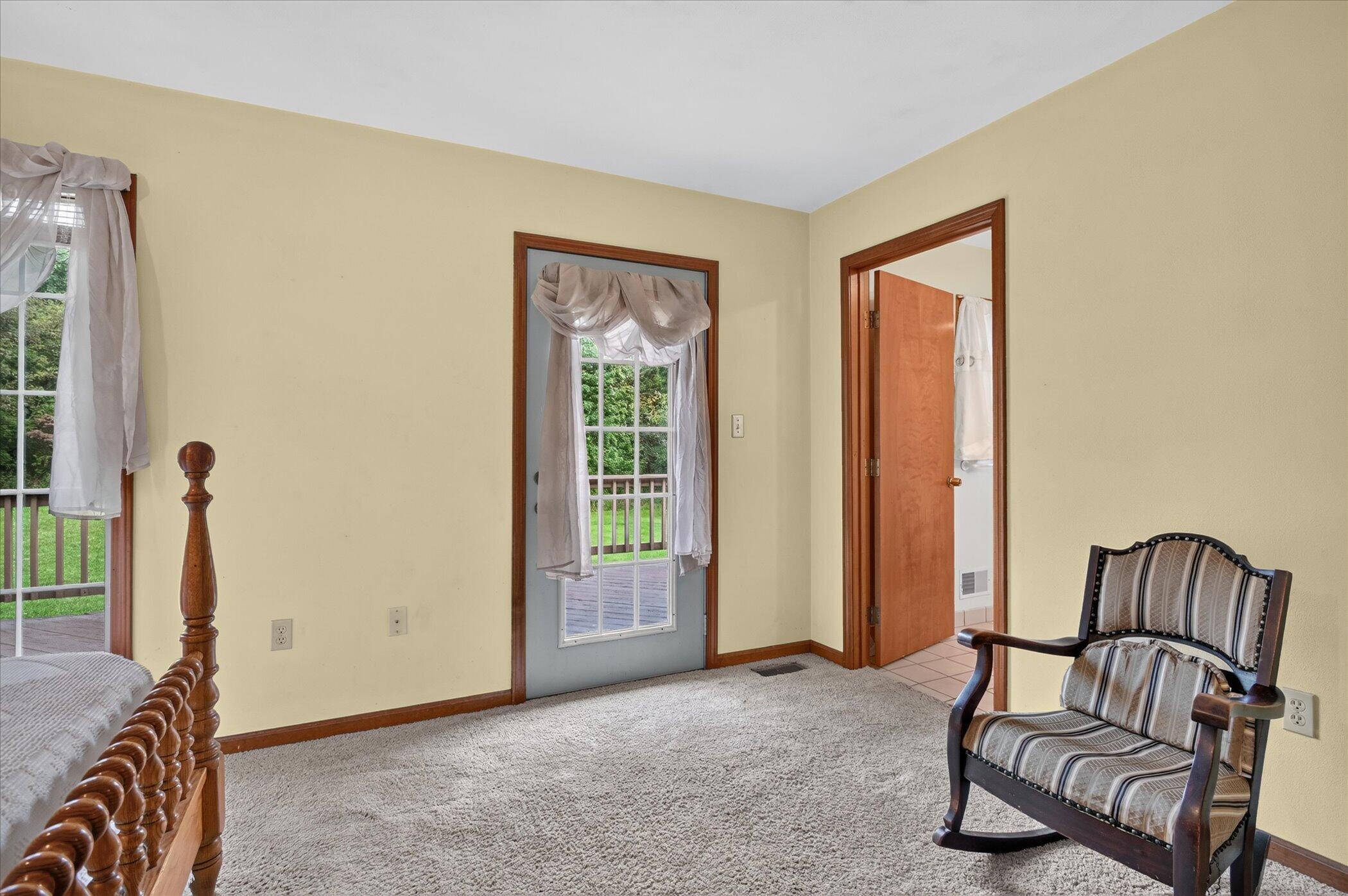 Bedroom with a wooden bed, upholstered rocking chair, and windows overlooking a deck and lawn.