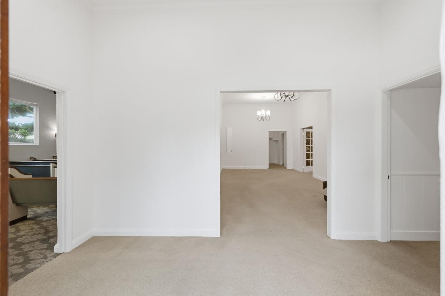 Carpeted room opening into a larger room with chandeliers and a glimpse of the kitchen.