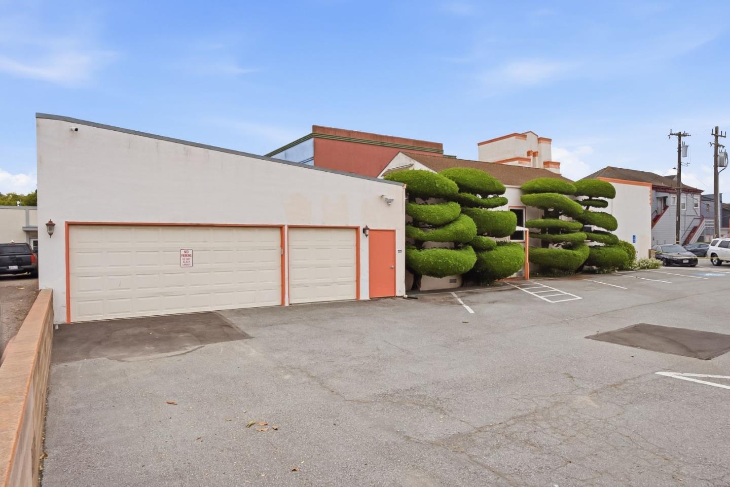 Building exterior with orange-trimmed garage doors and sculpted evergreen shrubs beside a parking area.