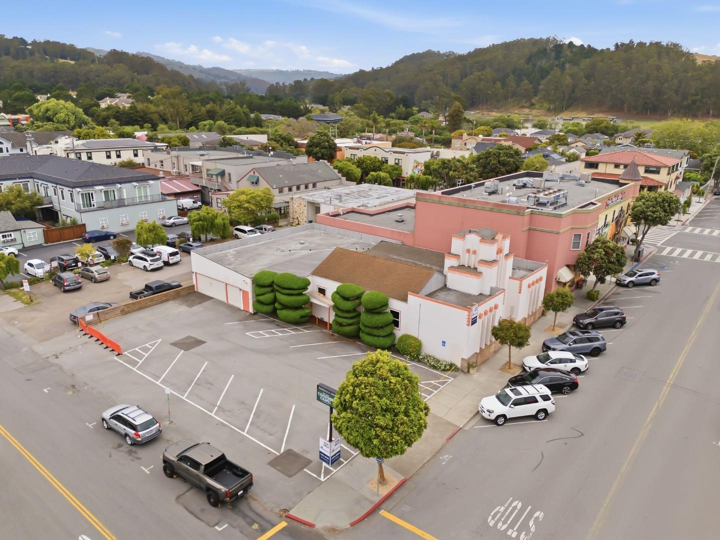 Aerial view of a commercial building and parking lot, with town buildings and wooded hills beyond.