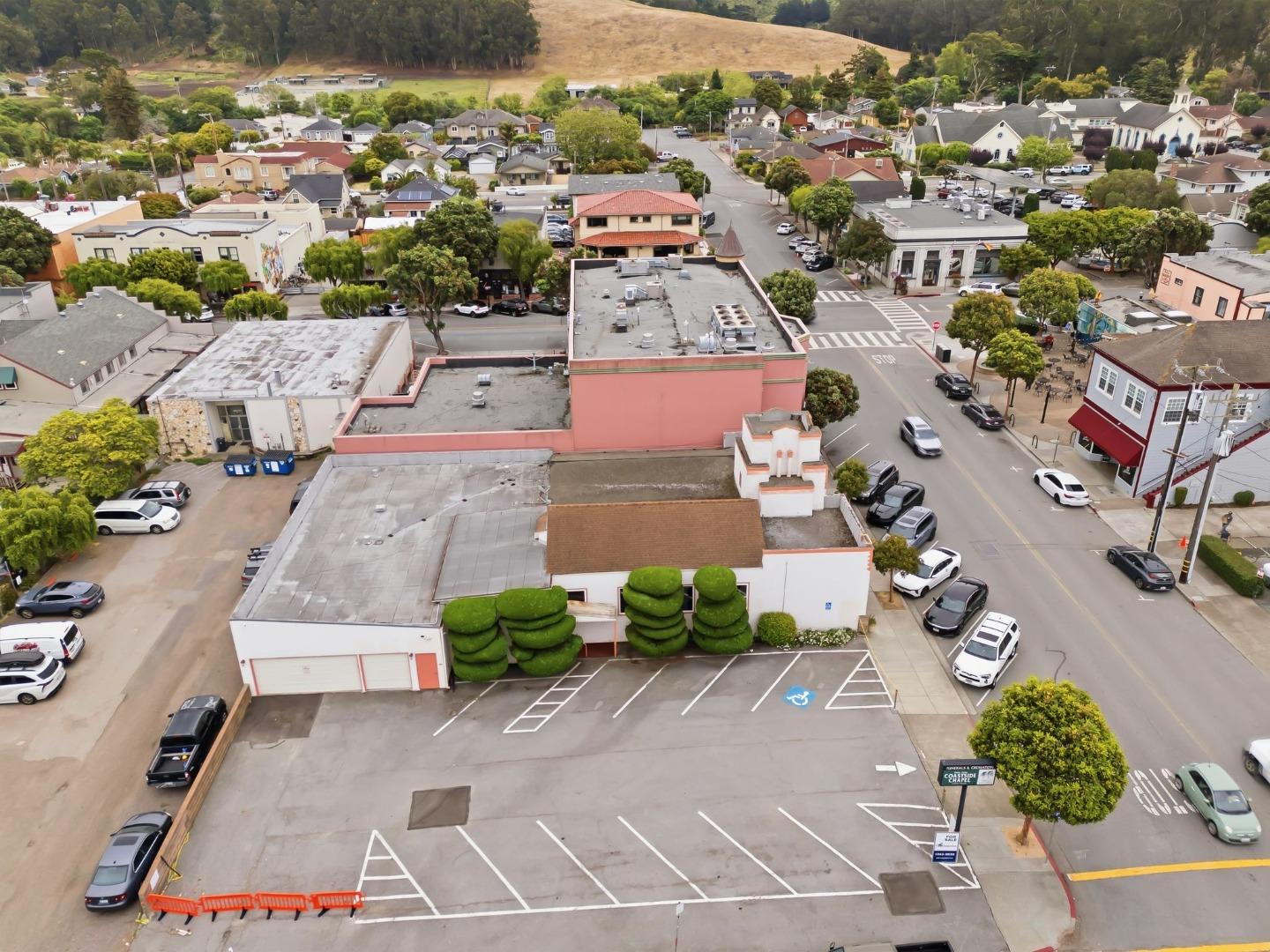 Aerial view of a commercial property and parking lot, with a town intersection and wooded hills beyond.