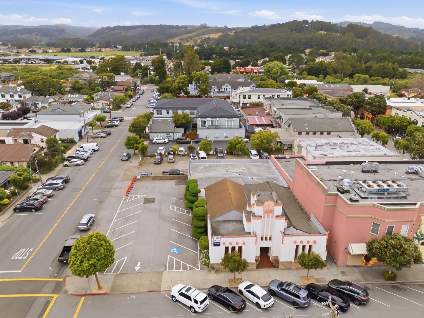 Aerial view of a Half Moon Bay neighborhood with parking lots, low-rise buildings, and wooded hills.