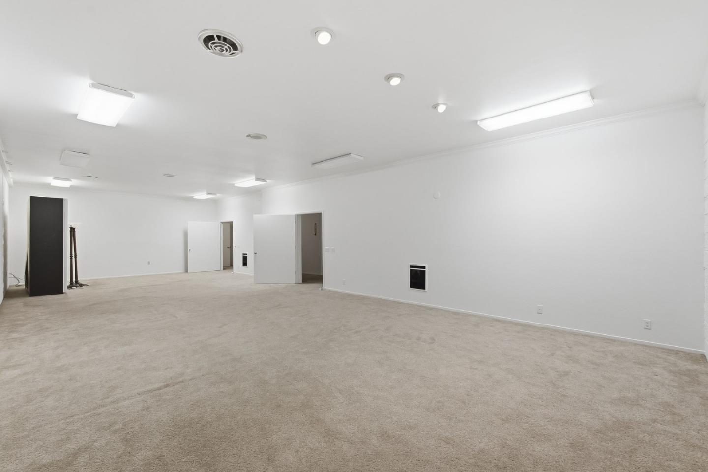 Large vacant carpeted room with white walls, multiple ceiling lights, and open doorways.