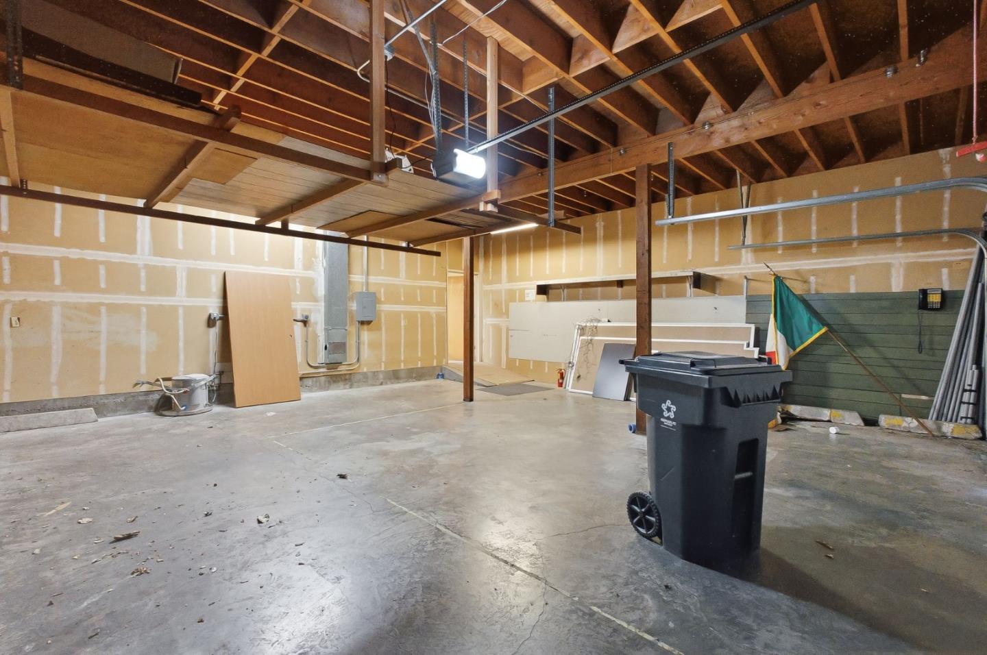 Vacant garage with a concrete floor, unfinished walls, exposed rafters, and a wheeled trash bin.
