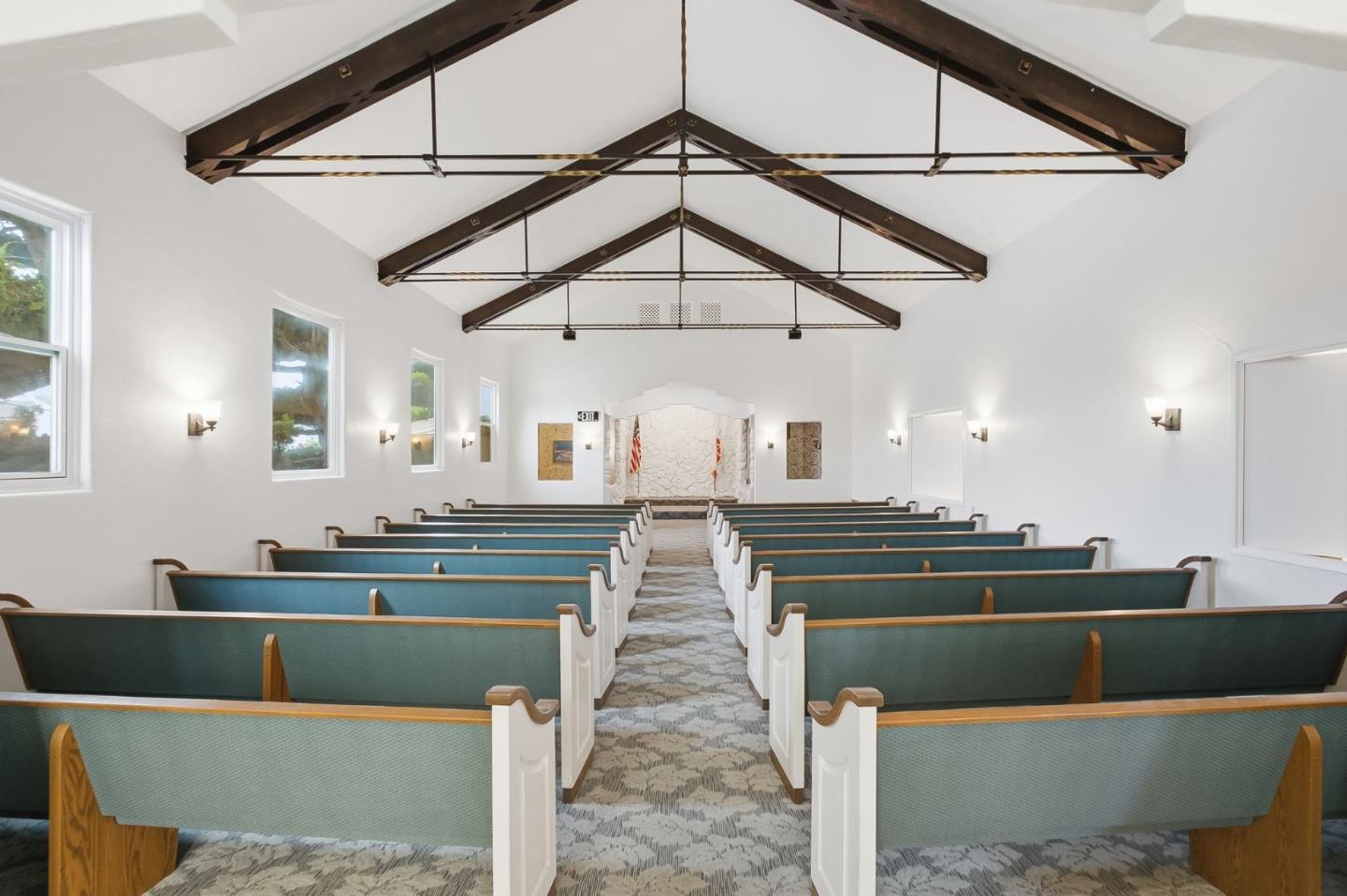 Chapel interior with teal pews, a patterned center aisle, and dark exposed ceiling trusses.