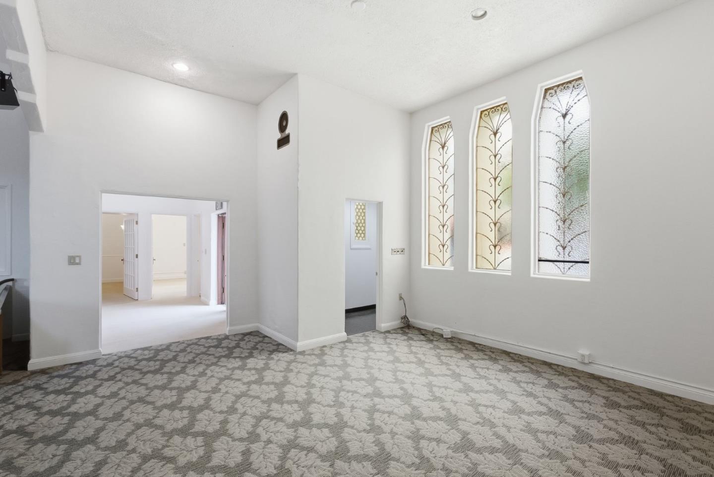 Empty room with patterned carpet, a high ceiling, and three tall arched decorative windows.