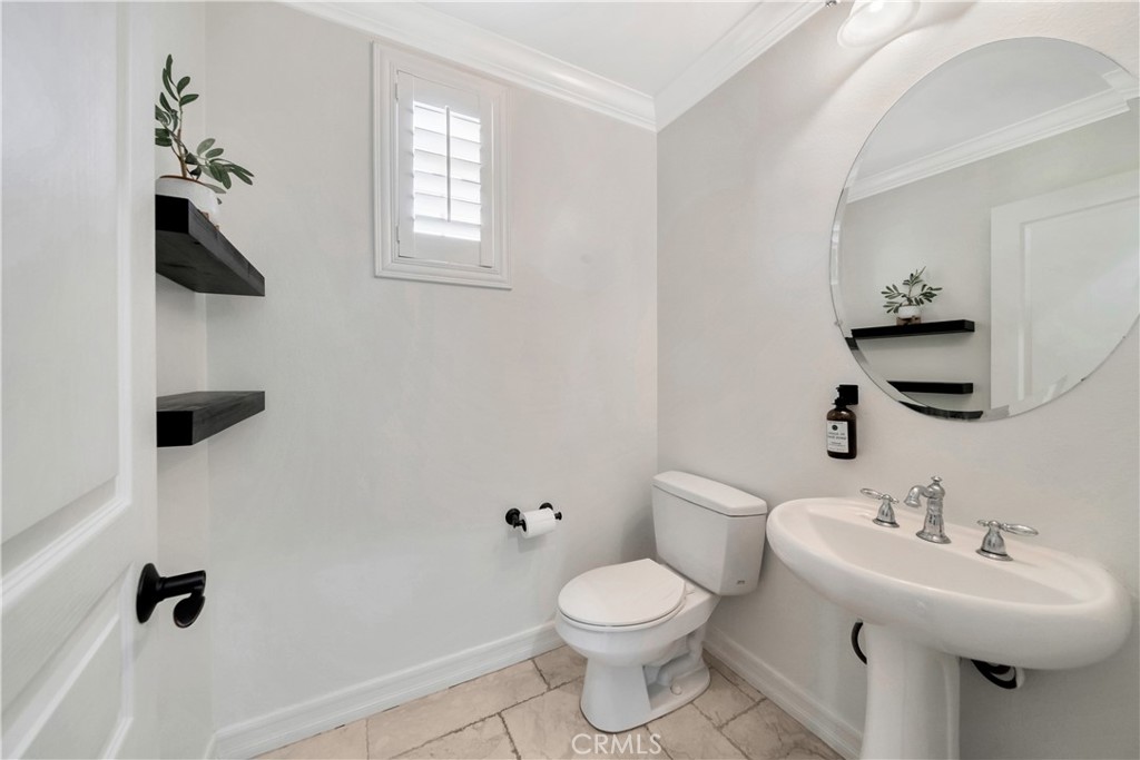 Bathroom with a pedestal sink, round mirror, black floating shelves, and marble-look tile floor.