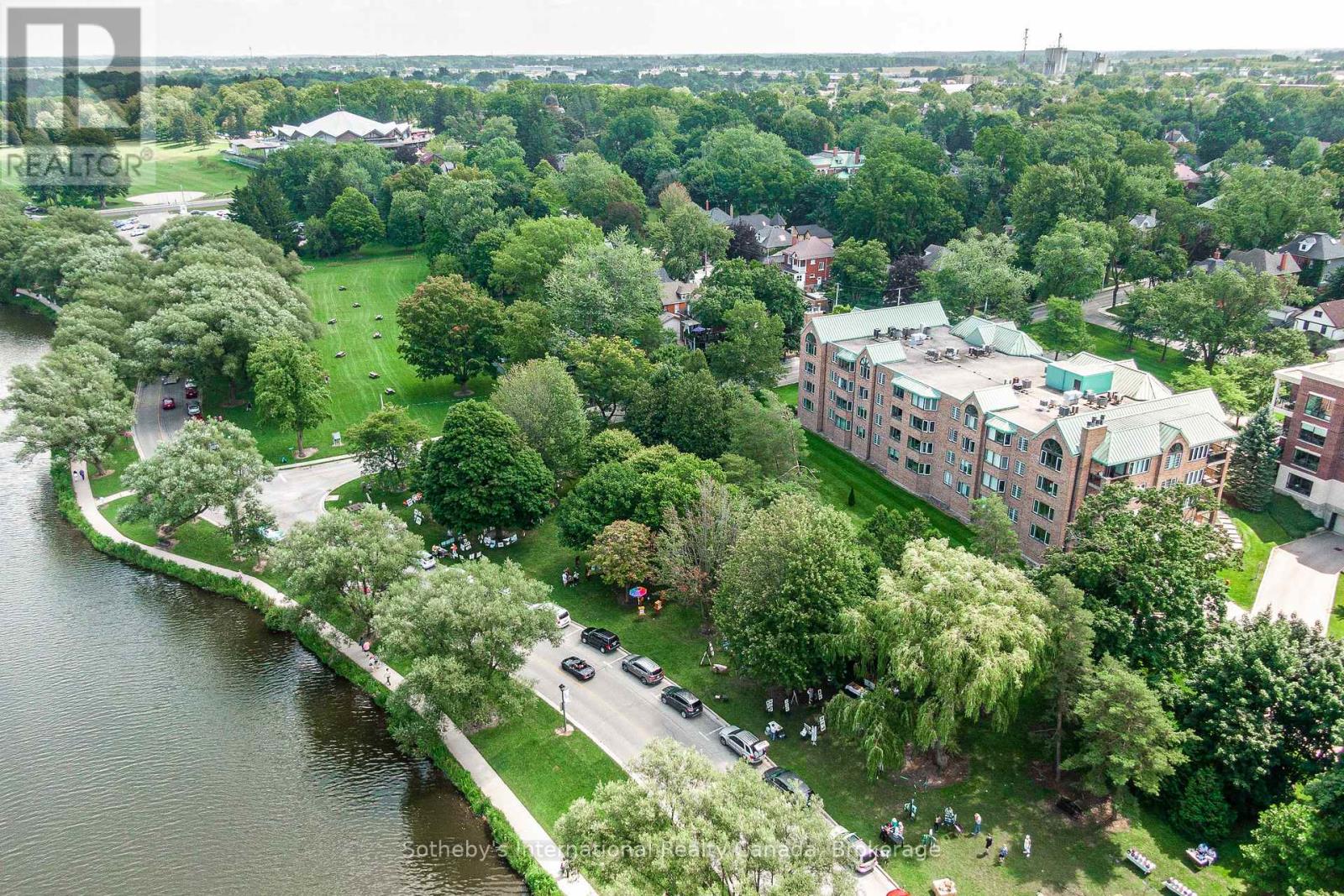 Aerial view of a brick condominium beside a river, park, and tree-lined waterfront path.