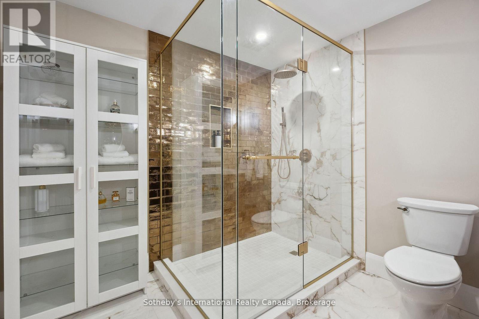 Bathroom with a gold-trimmed glass shower, bronze tile, marble-look surfaces, and white glass-front cabinets.