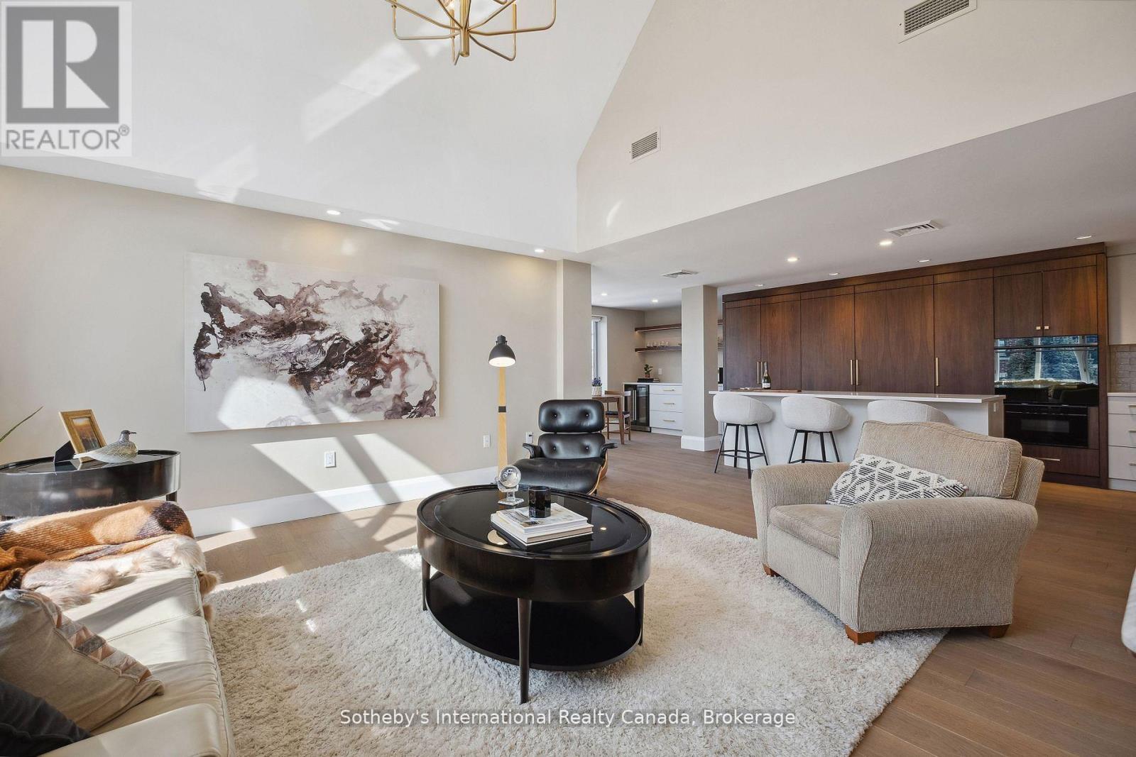 Open-plan condo living room with vaulted ceiling, abstract artwork, neutral seating, and a dark wood kitchen.