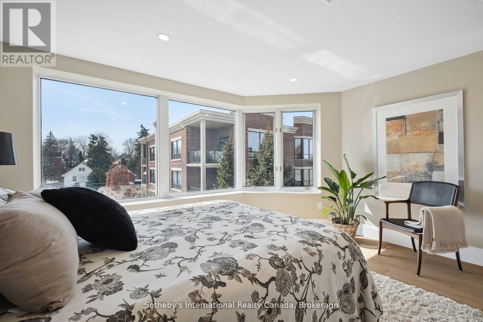Bedroom with a floral bedspread, corner windows overlooking nearby homes, and a chair beside a potted plant.