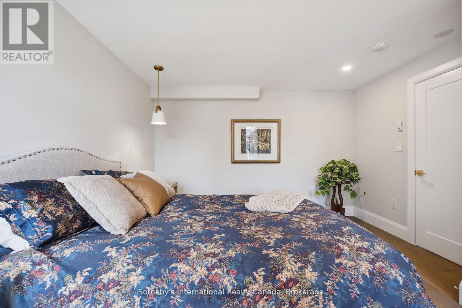 Furnished bedroom with blue floral bedding, a light upholstered headboard, framed artwork, and a potted plant.