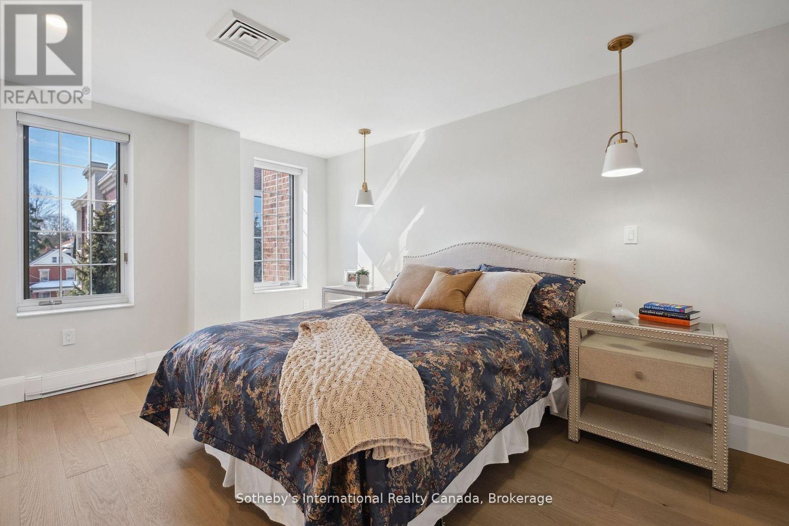 Staged bedroom with a floral-covered bed, two windows, and pendant lights.