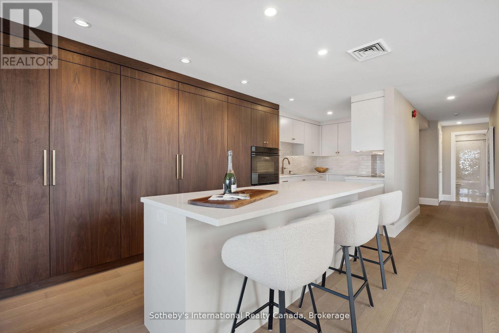 Modern kitchen with a large white island, three upholstered stools, and dark walnut cabinetry.