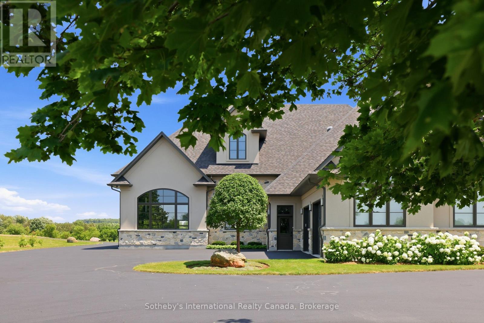 Beige house with a stone base, steep roof, arched window, and broad paved driveway.