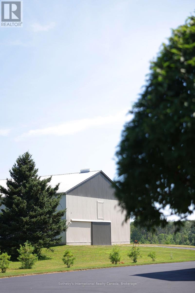 Gray barn-like outbuilding with a dark sliding door, lawn, evergreen trees, and wooded hills.