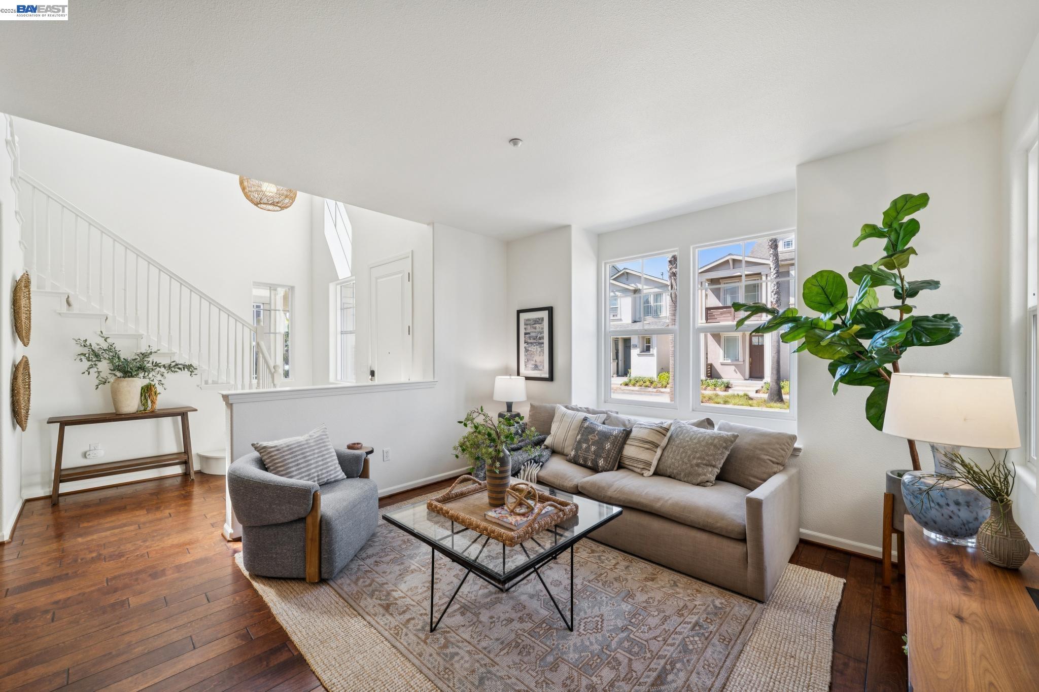 Staged living room with hardwood floors, a staircase, gray sofa, and large windows.
