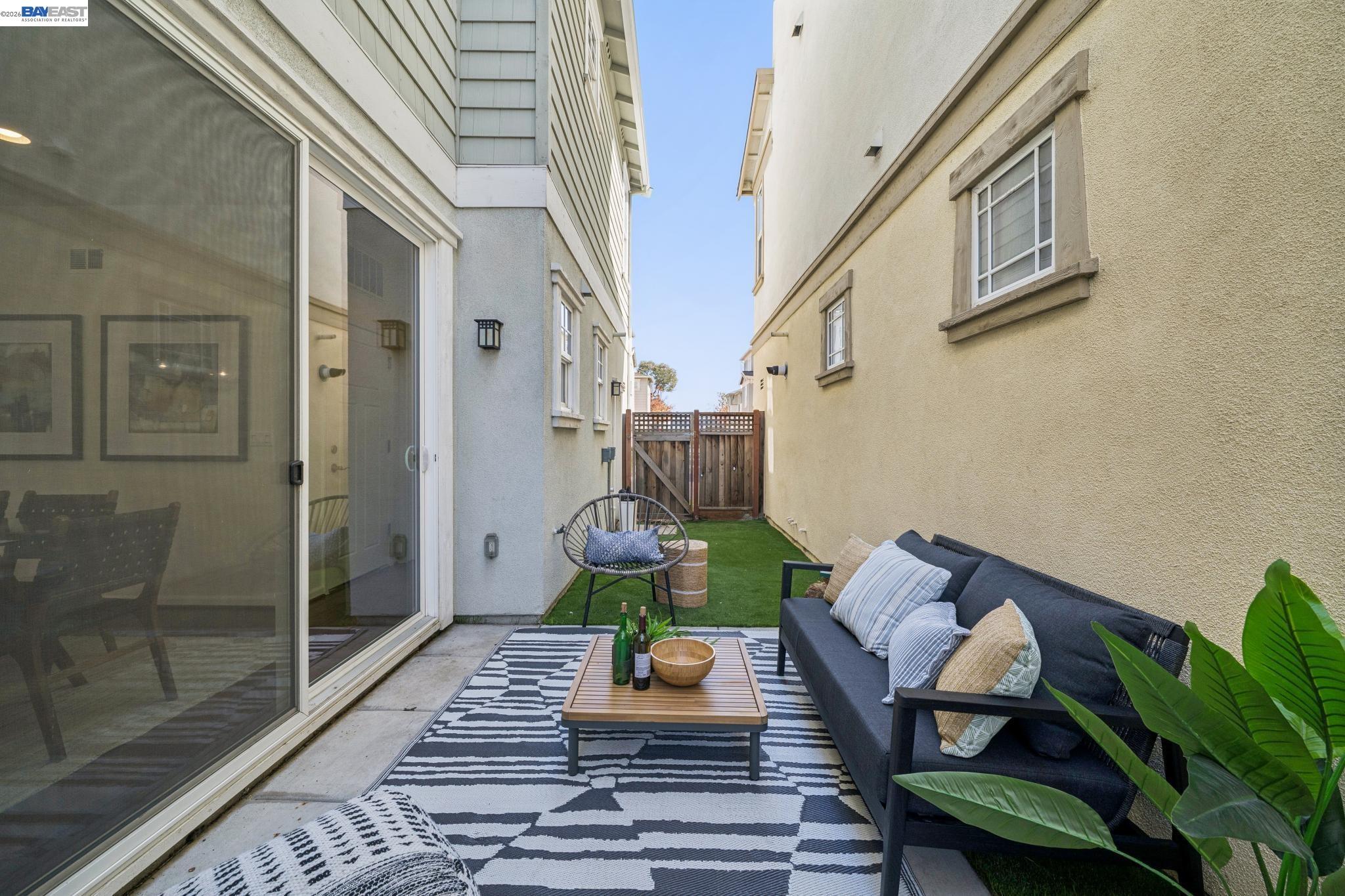 Narrow staged patio with artificial turf, outdoor sofa, striped rug, and wooden gate.