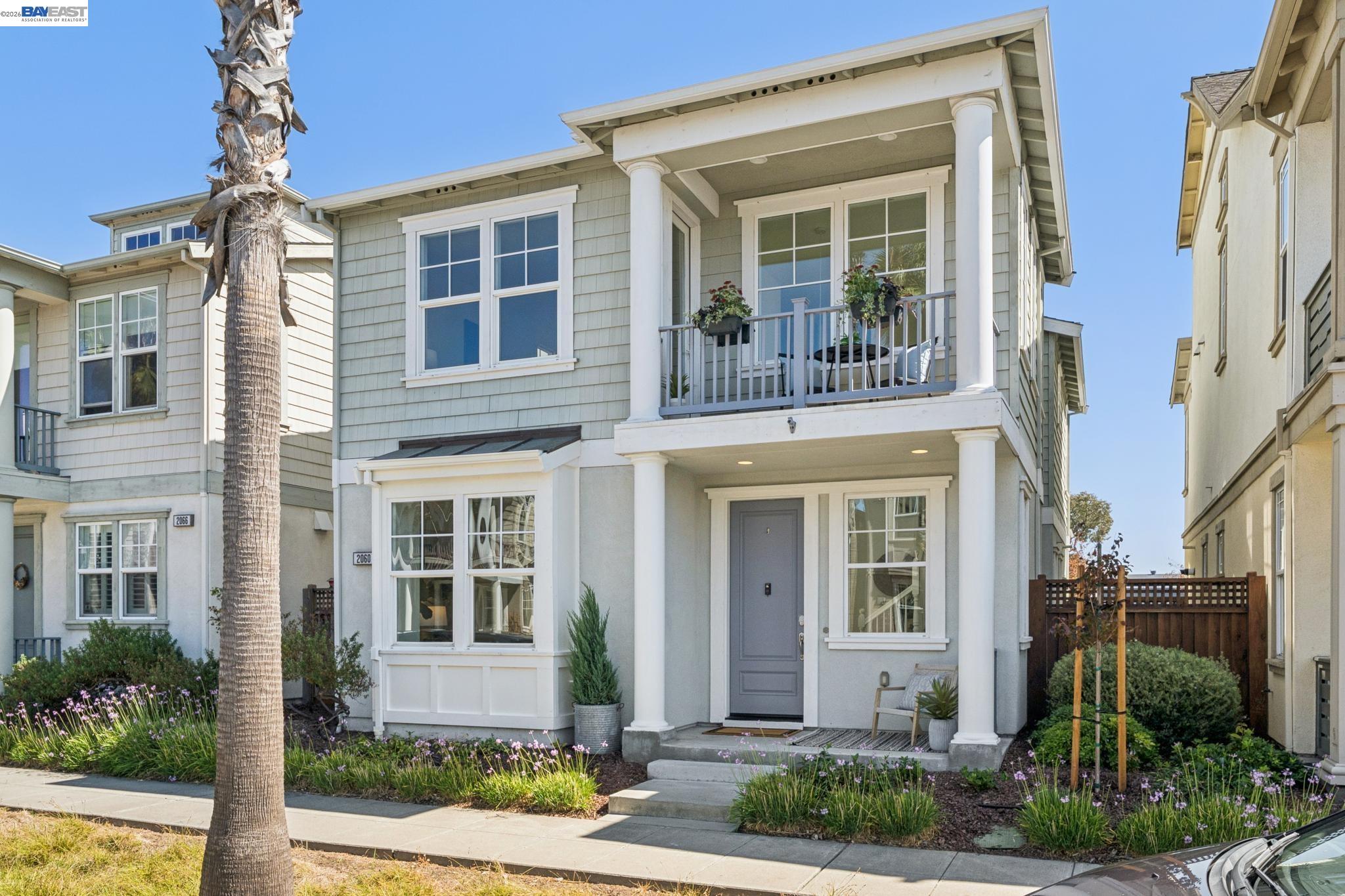 Two-story gray home with a columned porch, flower-filled balcony, and palm tree by the sidewalk.