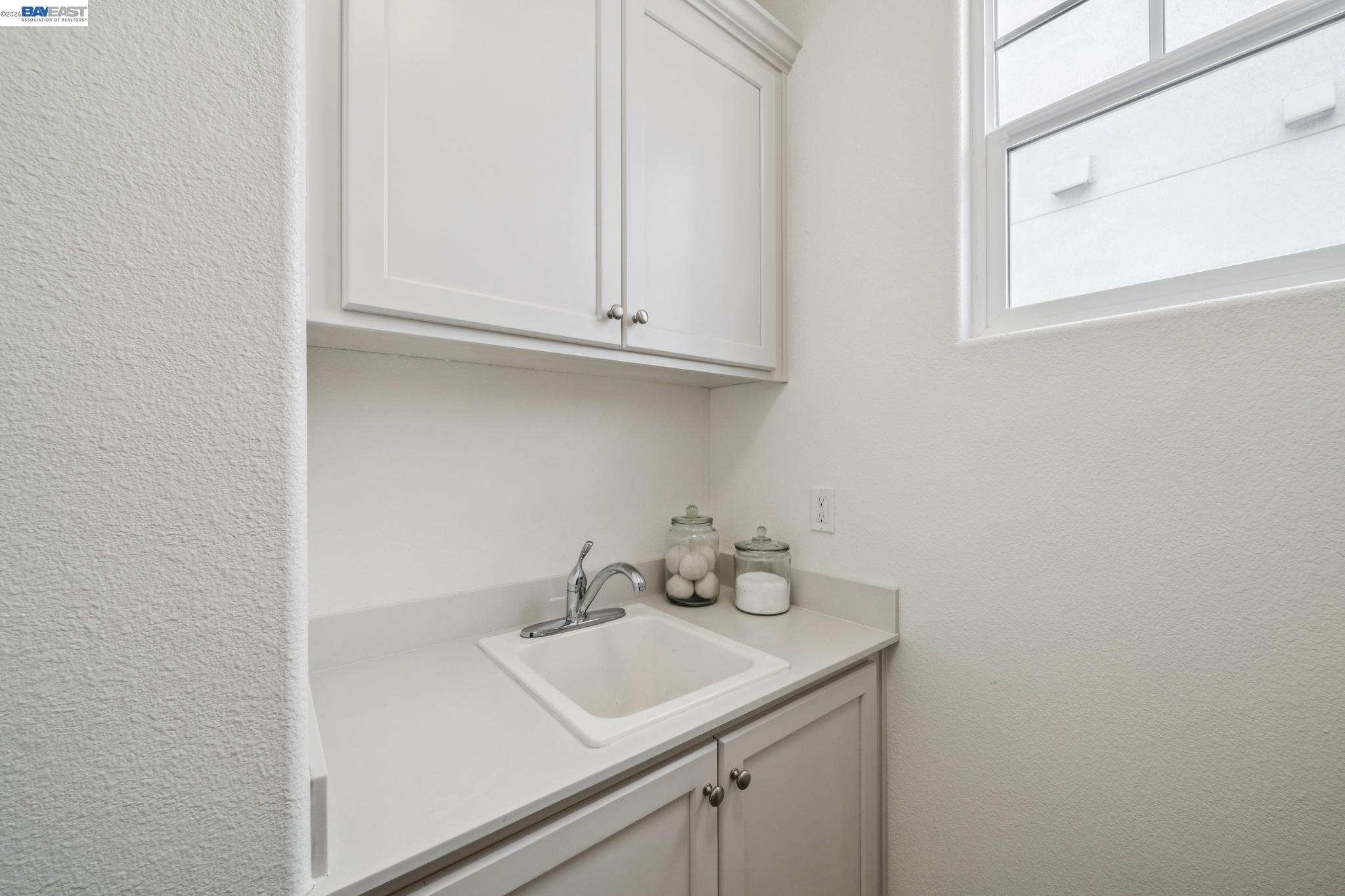 Utility sink with white cabinetry, chrome faucet, countertop jars, and a window.