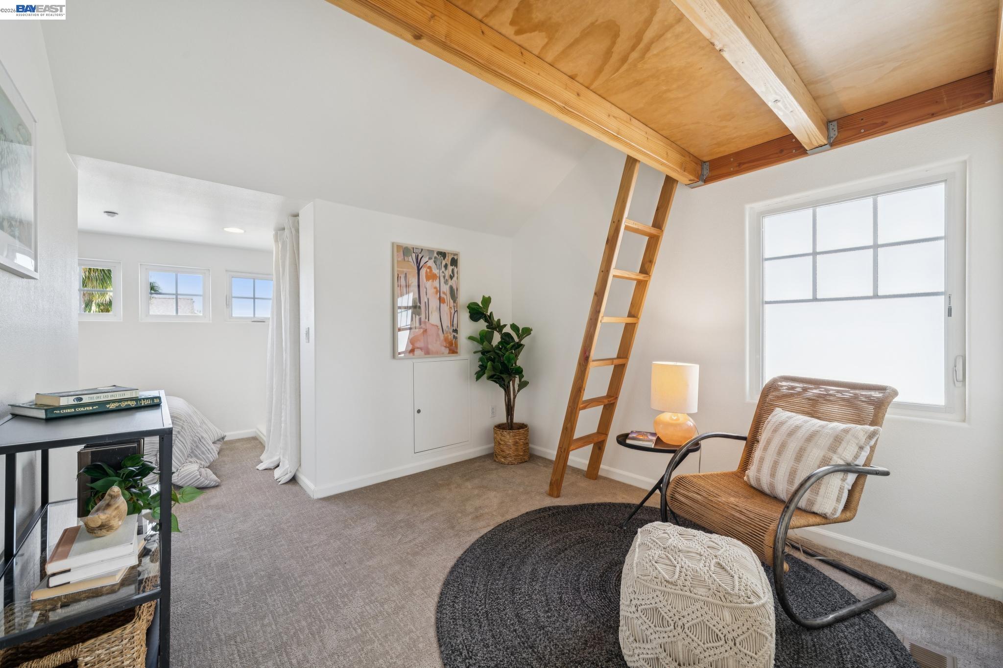 Staged loft bedroom with exposed wooden beams, a ladder, and a wicker chair beside a large window.