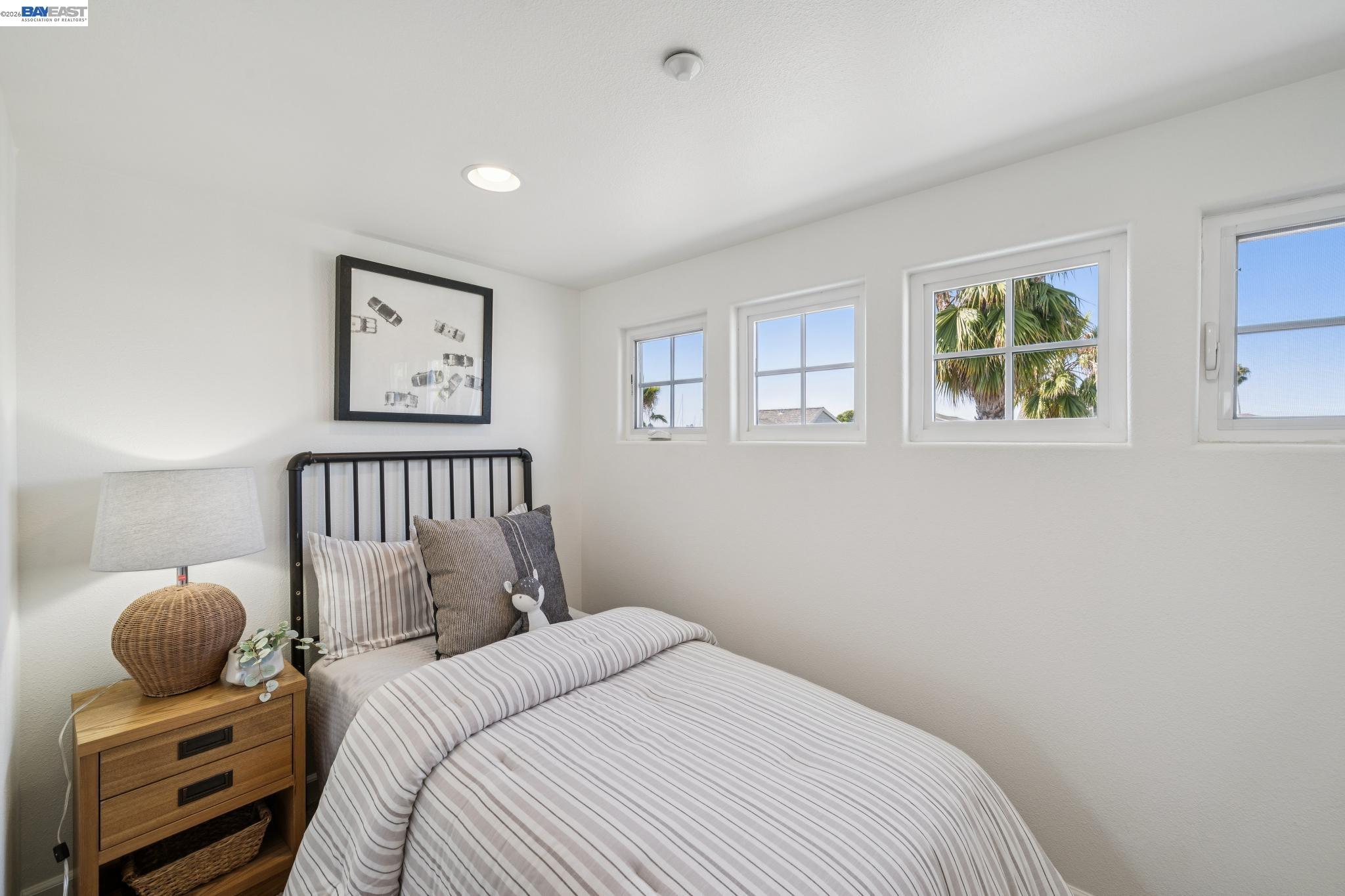 Furnished bedroom with striped bedding, a black metal headboard, and windows overlooking palm trees.
