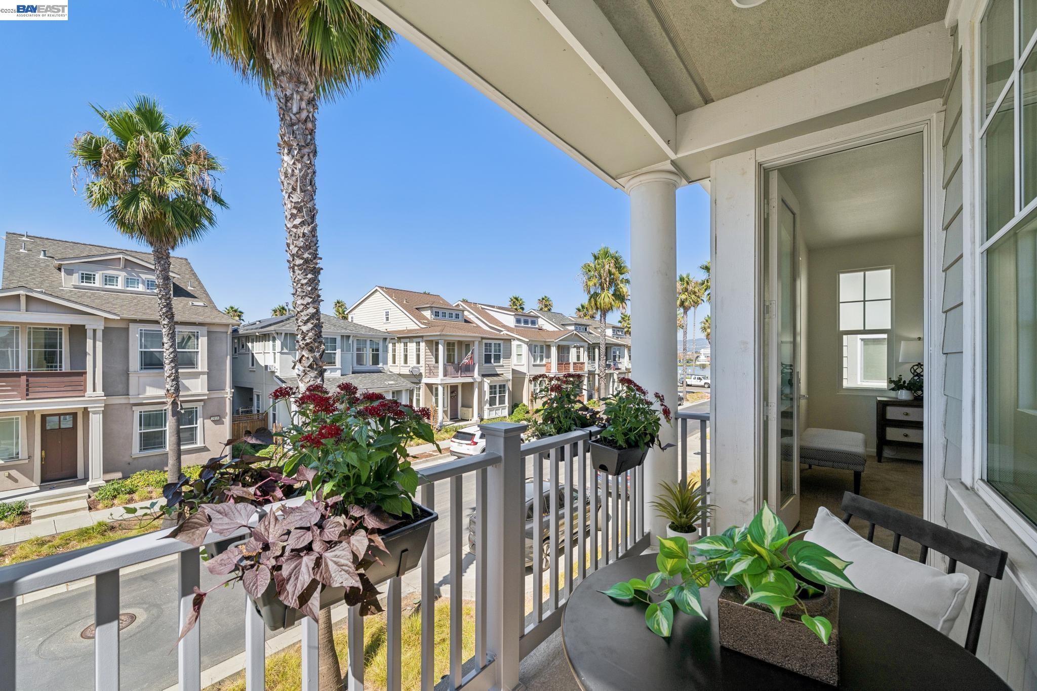 Covered balcony with potted plants and seating overlooking palm-lined townhomes.