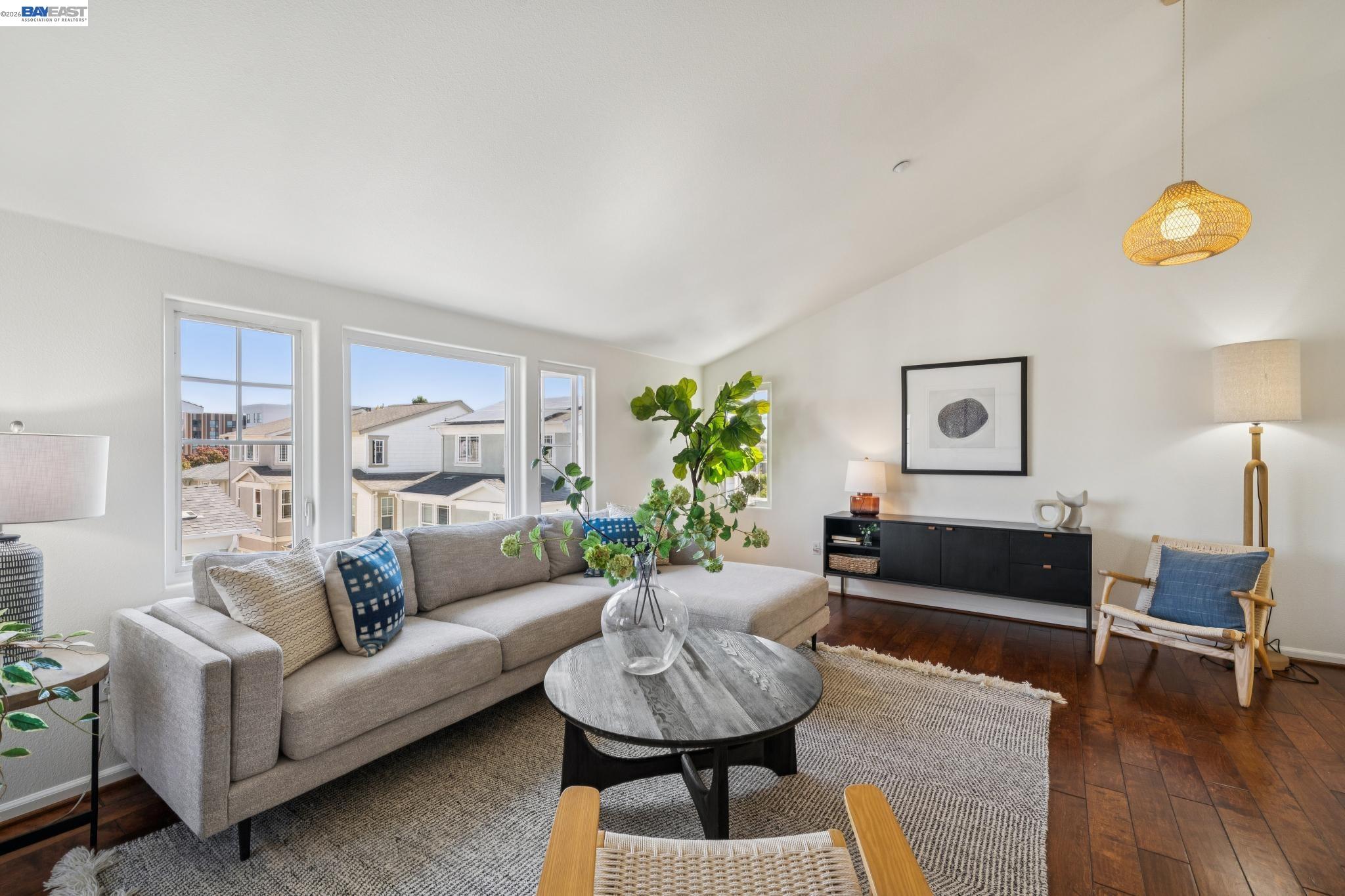 Staged living room with a gray sectional, round coffee table, dark wood floors, and large windows.