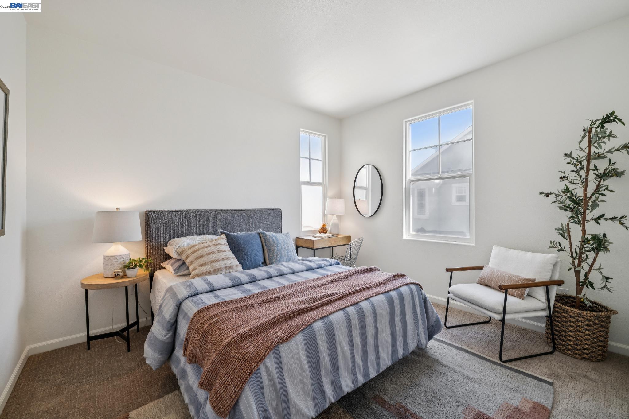 Staged bedroom with blue striped bedding, a rust knit throw, and a white accent chair beside two windows.