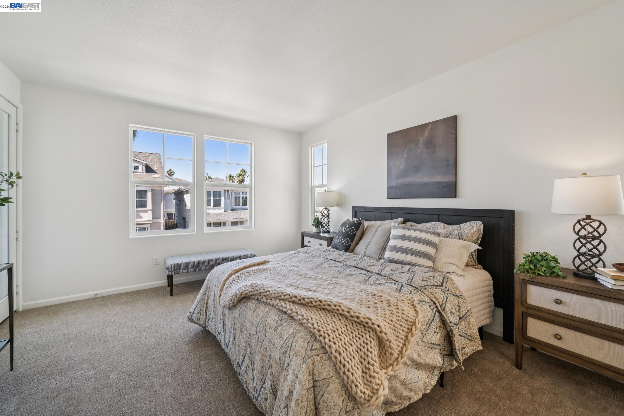 Furnished bedroom with patterned bedding, a chunky knit throw, and windows overlooking neighboring homes.